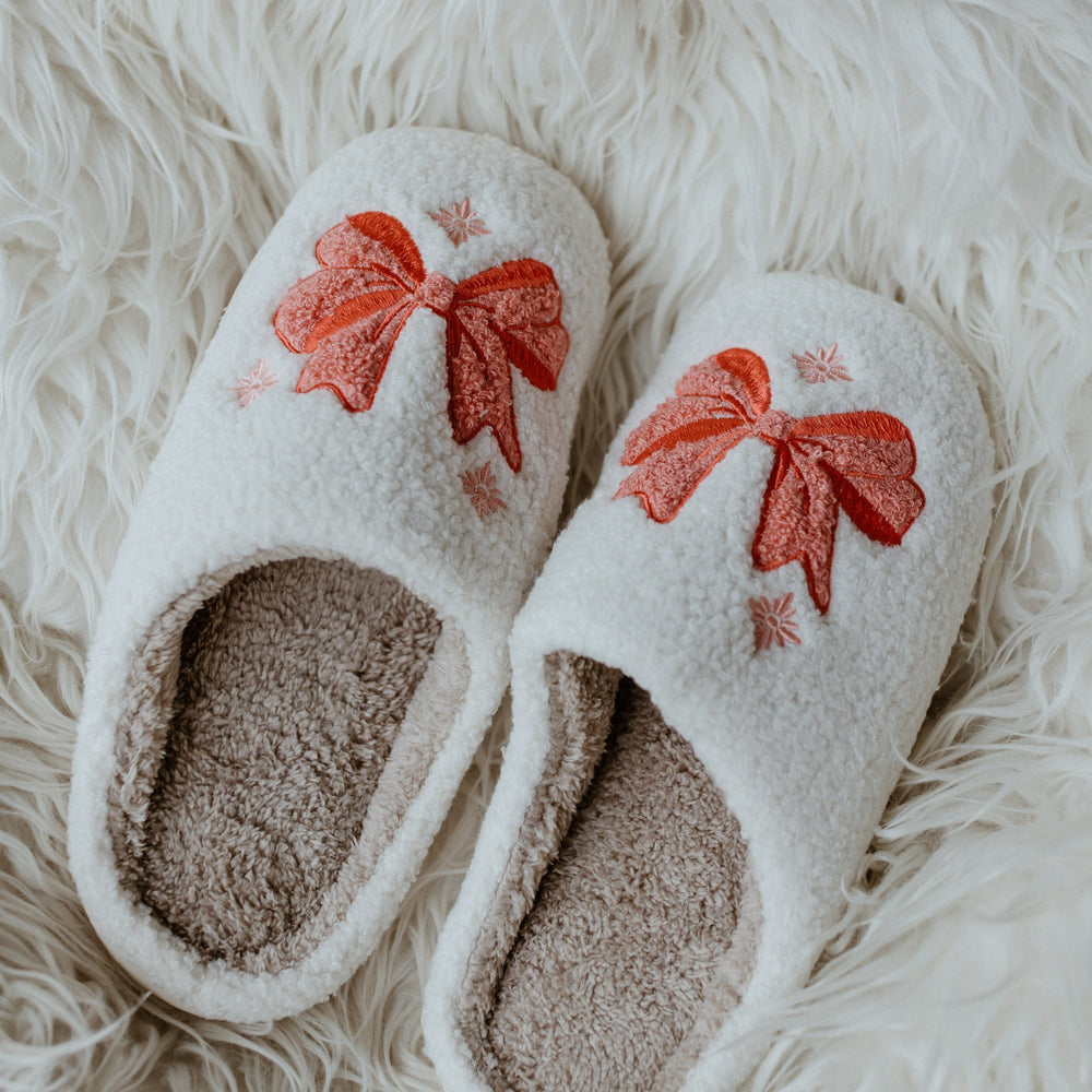 White holiday bow fuzzy slippers with red bow designs, placed on a fluffy white surface, add a touch of festive charm to any cozy setting.
