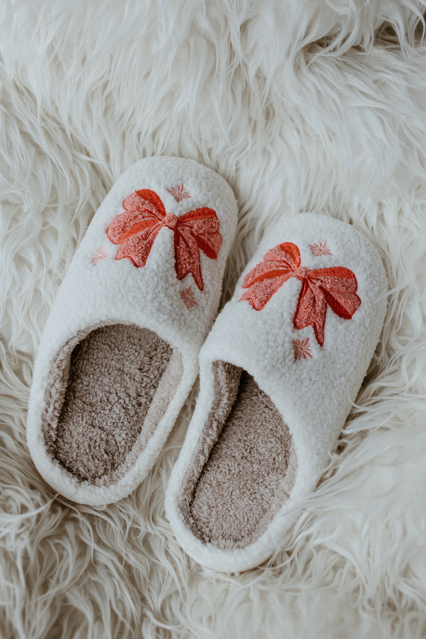 White holiday bow fuzzy slippers with red bow designs, placed on a fluffy white surface, add a touch of festive charm to any cozy setting.