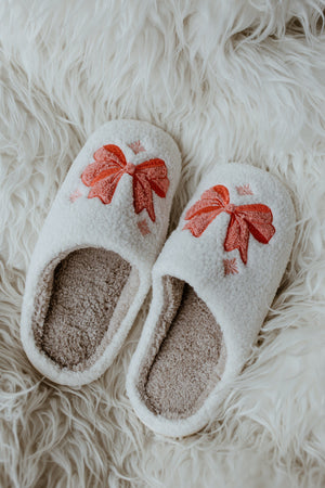 White holiday bow fuzzy slippers with red bow designs, placed on a fluffy white surface, add a touch of festive charm to any cozy setting.