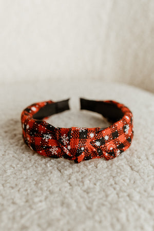 A red and black plaid holiday headband with a knotted design and white snowflake patterns rests on a soft, textured surface, offering an effortless style with a playful twist.