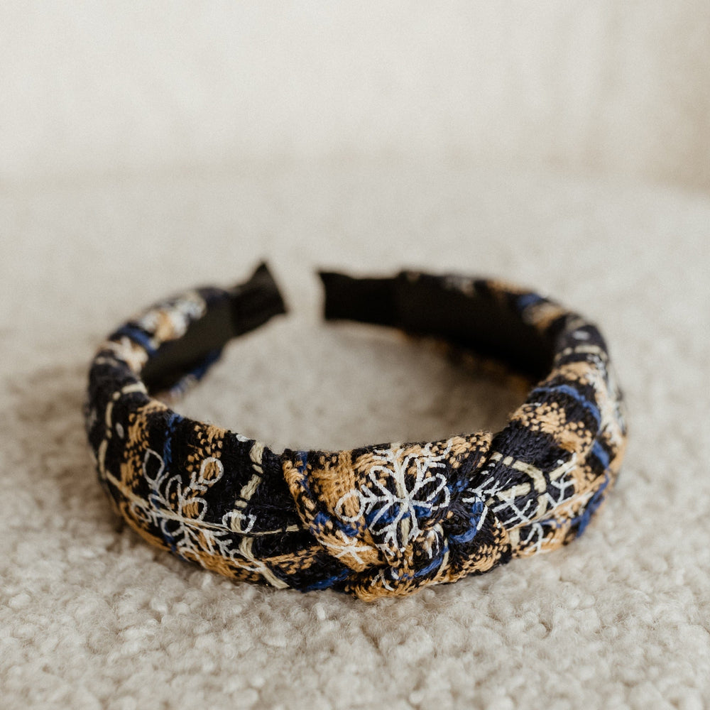 A black, blue, and gold floral embroidered holiday headband sits elegantly on a textured beige surface, perfect for completing your festive outfit.