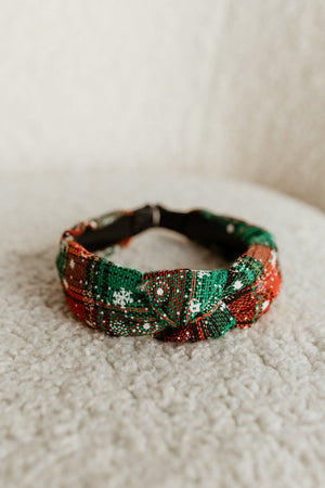 A festive polyester headband adorned with a red, green, and white plaid pattern lies elegantly on a textured light-colored surface, perfect as a holiday headband.