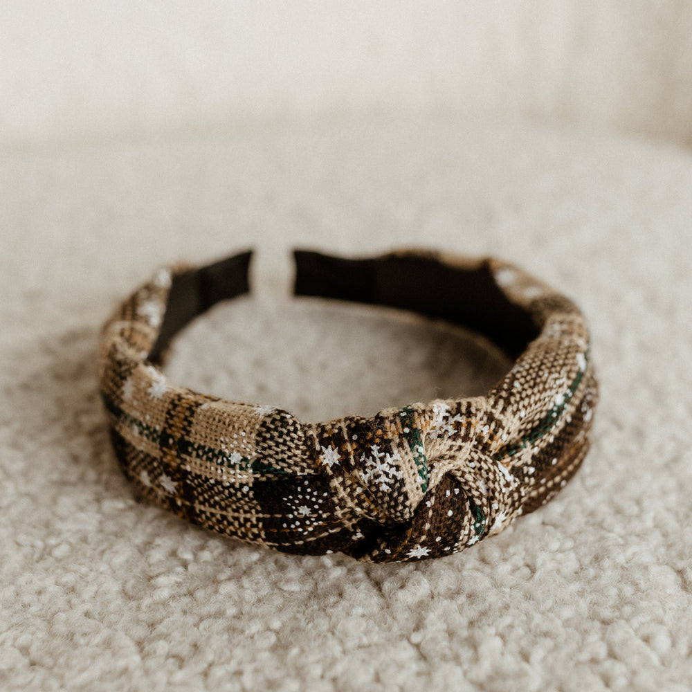 A plaid fabric headband with a knotted detail, perfect for completing your holiday outfit, lies on a soft, textured surface.
