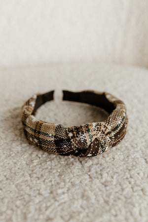 A plaid fabric headband with a knotted detail, perfect for completing your holiday outfit, lies on a soft, textured surface.