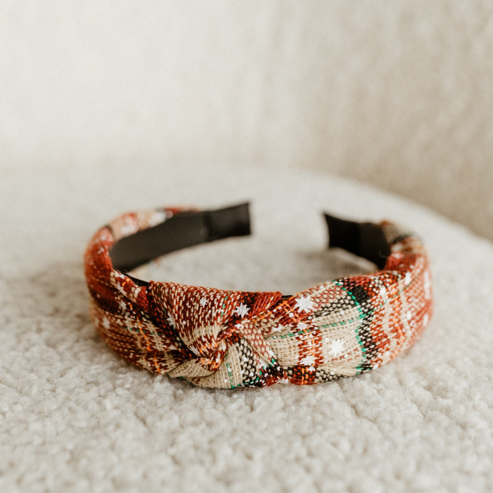 A holiday headband featuring a knotted design and tartan plaid pattern in red, beige, and green tones lies on a soft, textured surface. Crafted from polyester, it adds a touch of elegance to any festive ensemble.