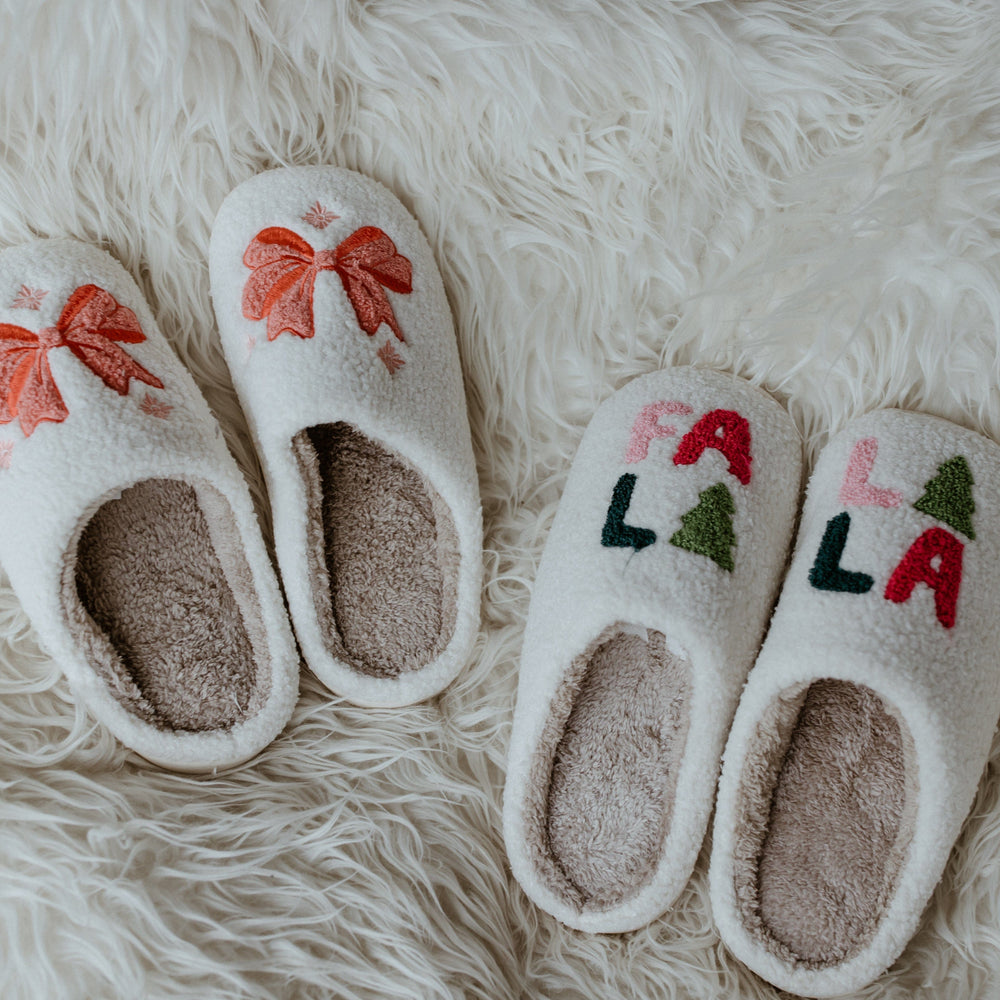 Two pairs of cozy slippers rest on a fluffy white rug. One pair features festive red bows, while the other displays "FA LA LA" with green trees, making this festive footwear perfect for the holiday season.