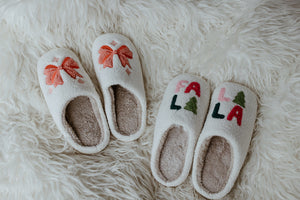 Two pairs of cozy slippers rest on a fluffy white rug. One pair features festive red bows, while the other displays "FA LA LA" with green trees, making this festive footwear perfect for the holiday season.