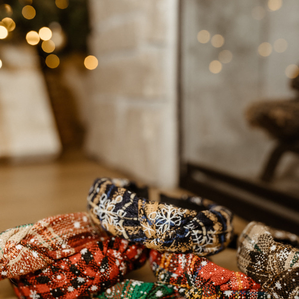 A collection of vibrant, polyester headbands adorned with holiday patterns, including a charming maple leaf design, arranged on a wooden surface with a softly blurred background of twinkling lights and a tree.