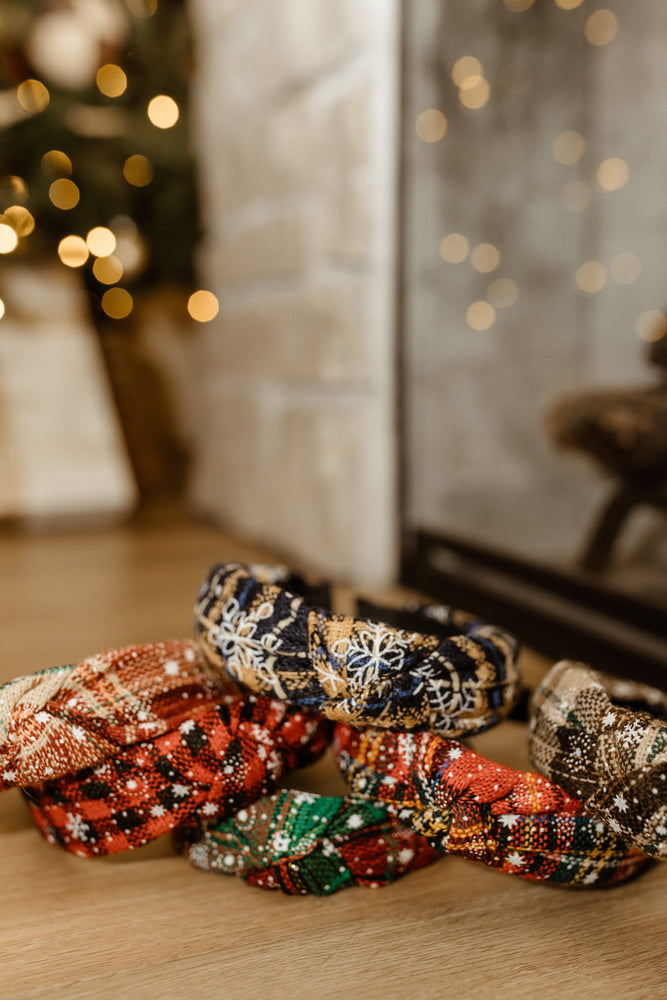 A collection of vibrant, polyester headbands adorned with holiday patterns, including a charming maple leaf design, arranged on a wooden surface with a softly blurred background of twinkling lights and a tree.