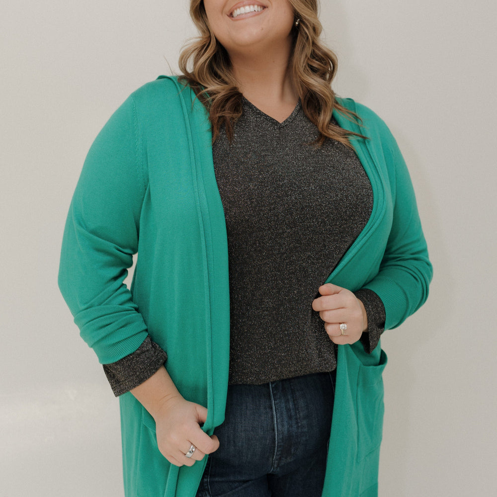 Woman wearing a teal cardigan over a dark shirt and jeans on a plain background