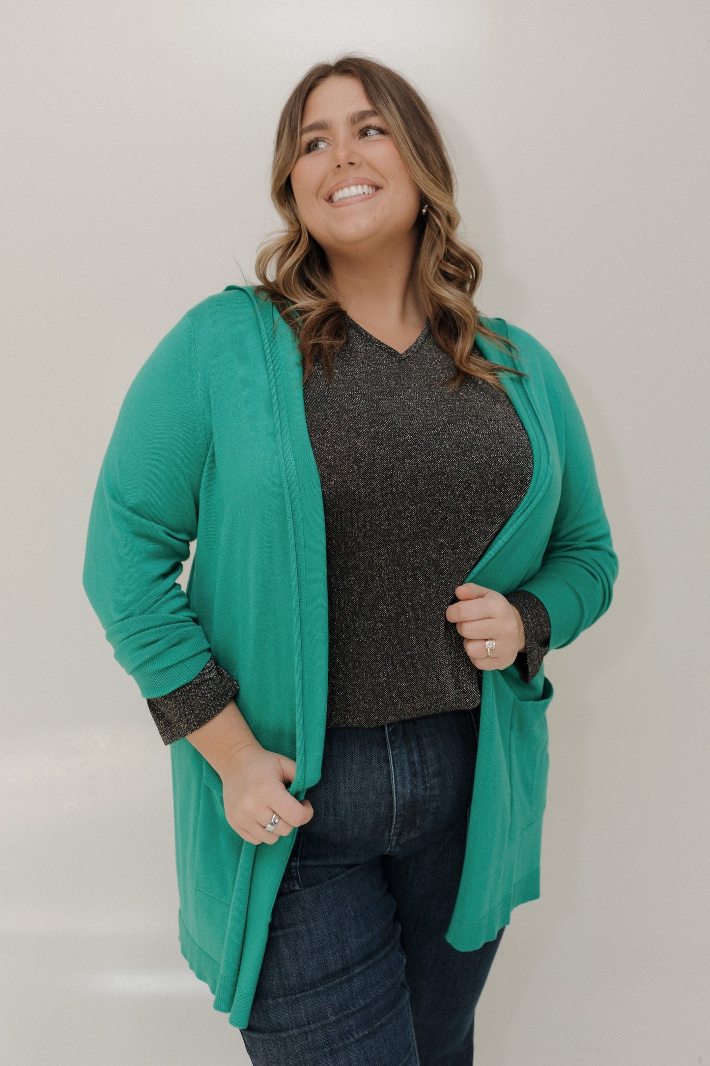Woman wearing a teal cardigan over a dark shirt and jeans on a plain background