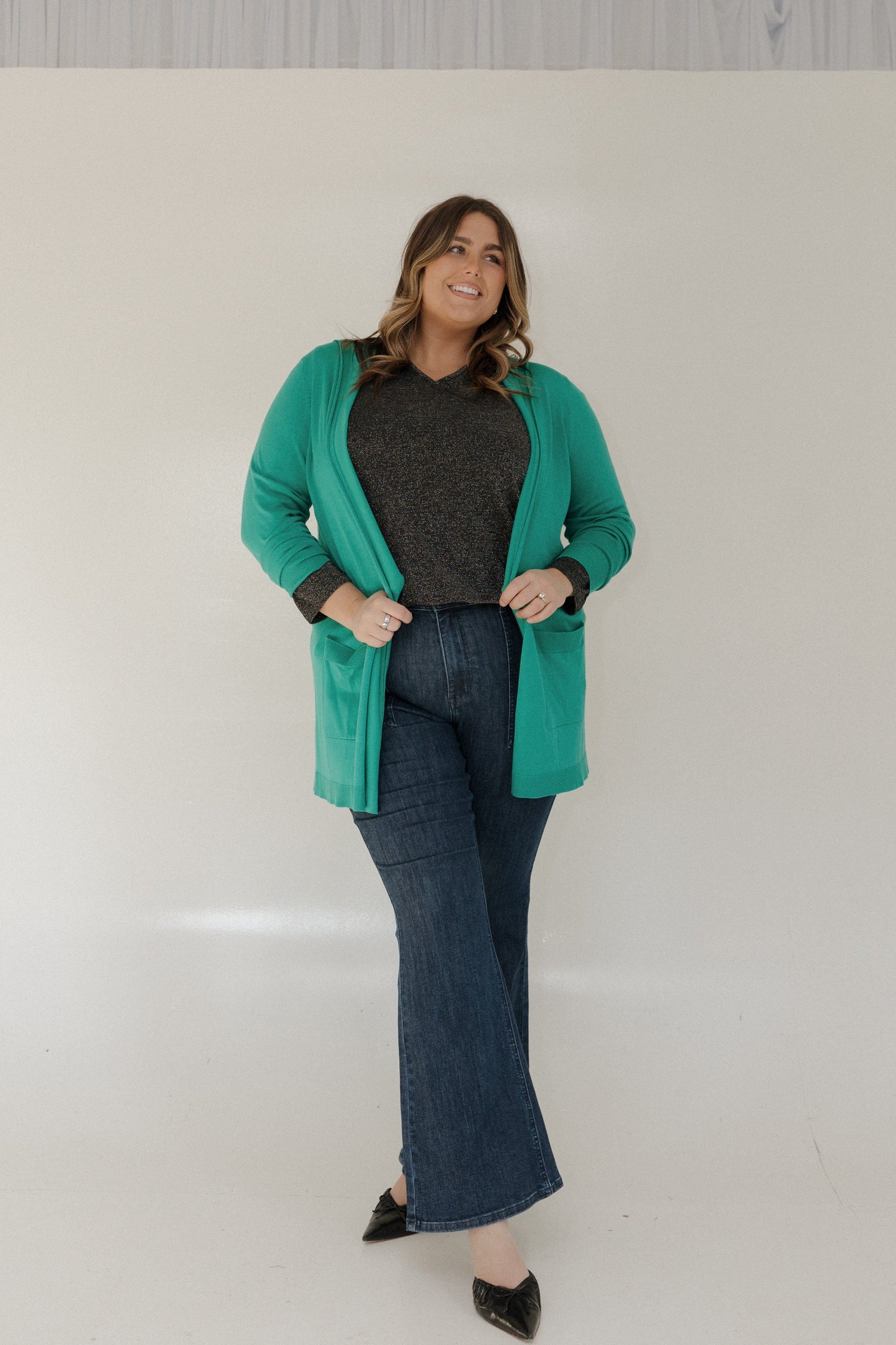 Woman wearing a green cardigan, black top, and blue jeans against a white background
