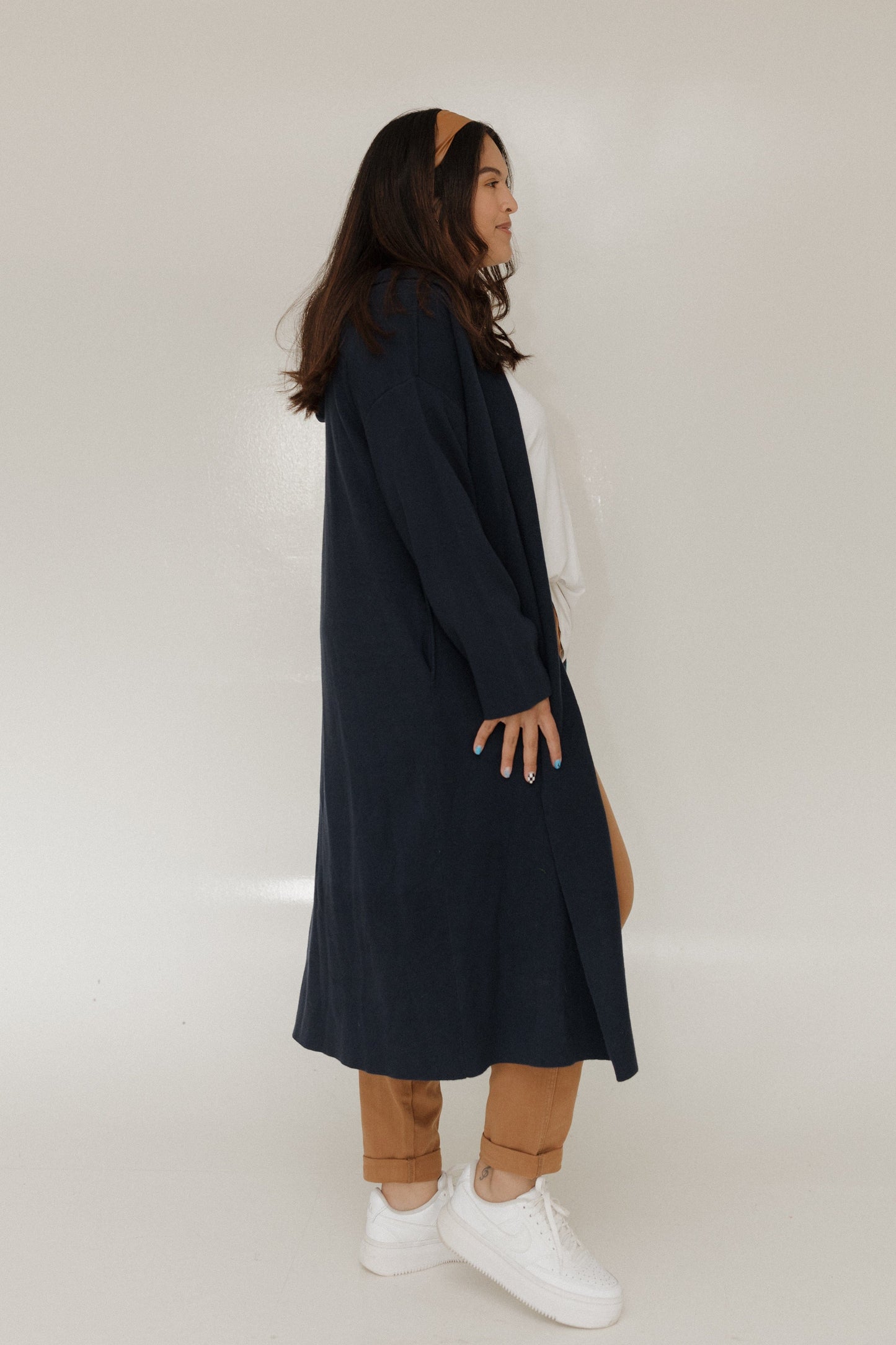 Person wearing a long dark coat over a white shirt with brown pants on a plain background