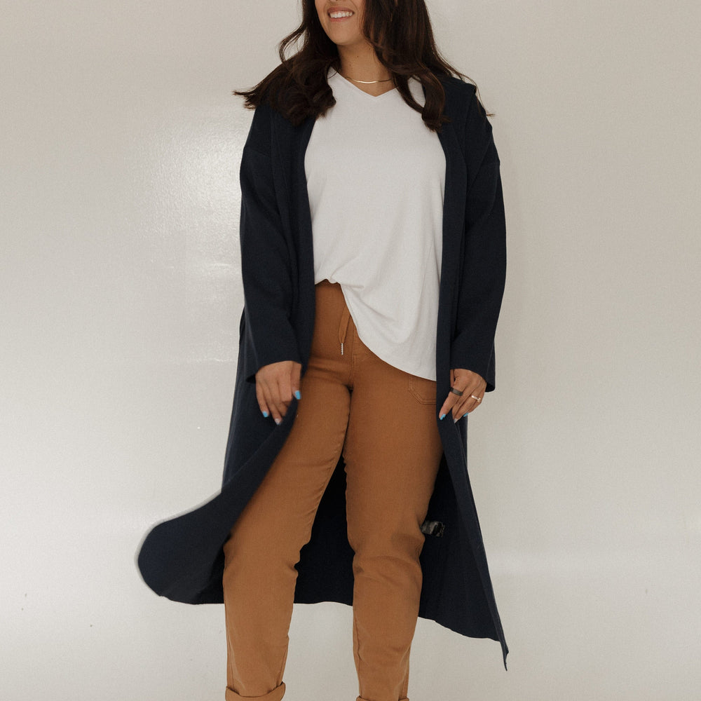 Woman wearing a long dark coat over a white top and brown pants on a plain background