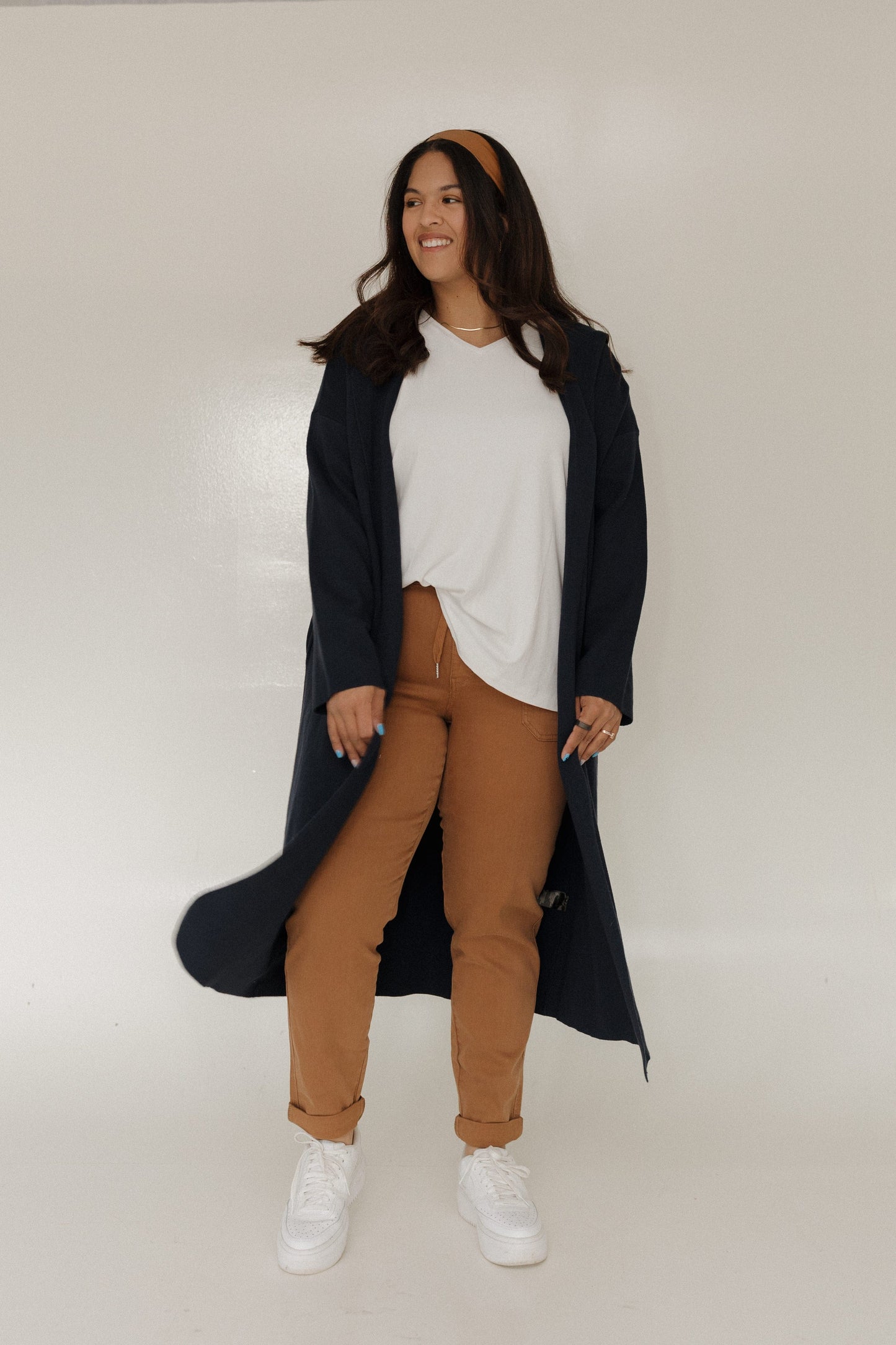 Woman wearing a long dark coat over a white top and brown pants on a plain background