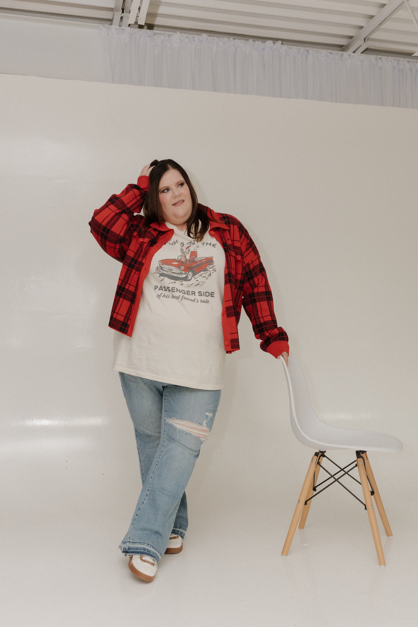 Person wearing a red plaid jacket over a graphic t-shirt with jeans, standing next to a white chair against a plain background.