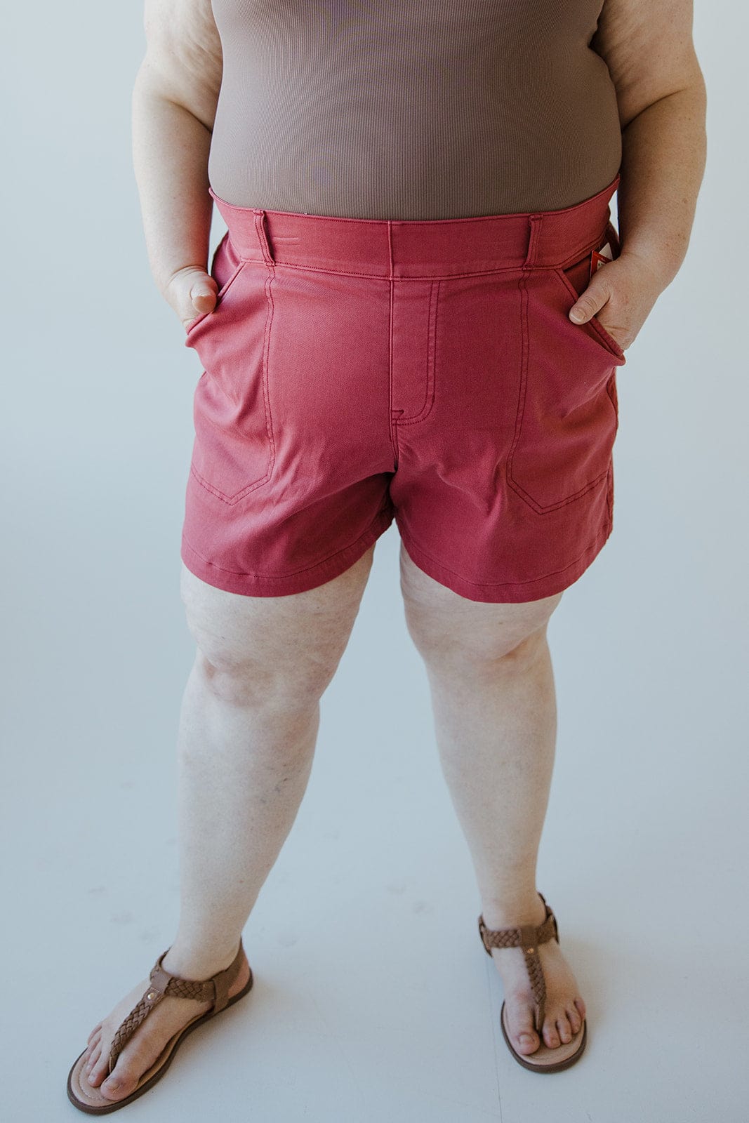 Person wearing a buttery-soft twill fabric sleeveless brown top, Spanx® STRETCH TWILL SHORTS 6" IN WILD ROSE, and sandals stands with their hands in their pockets against a plain background.