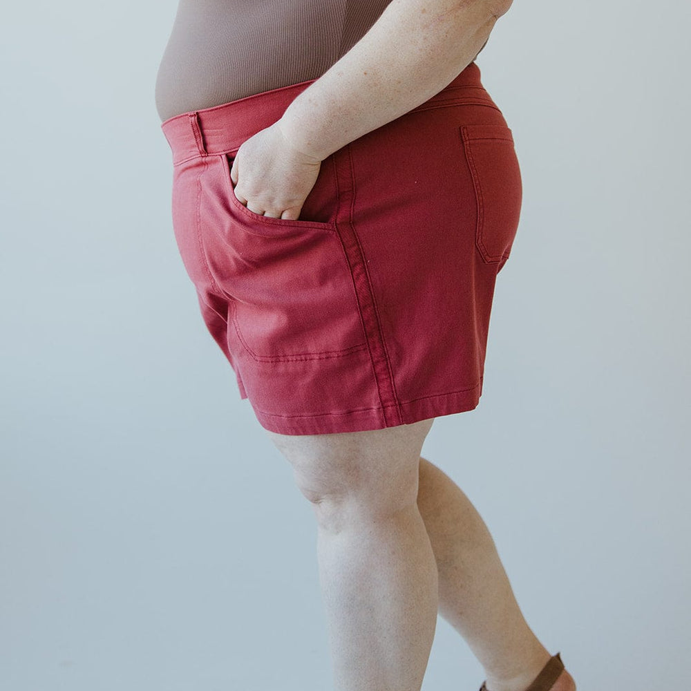 Person in a brown sleeveless top and Spanx® STRETCH TWILL SHORTS 6" IN WILD ROSE, with hands in pockets, standing against a plain background.