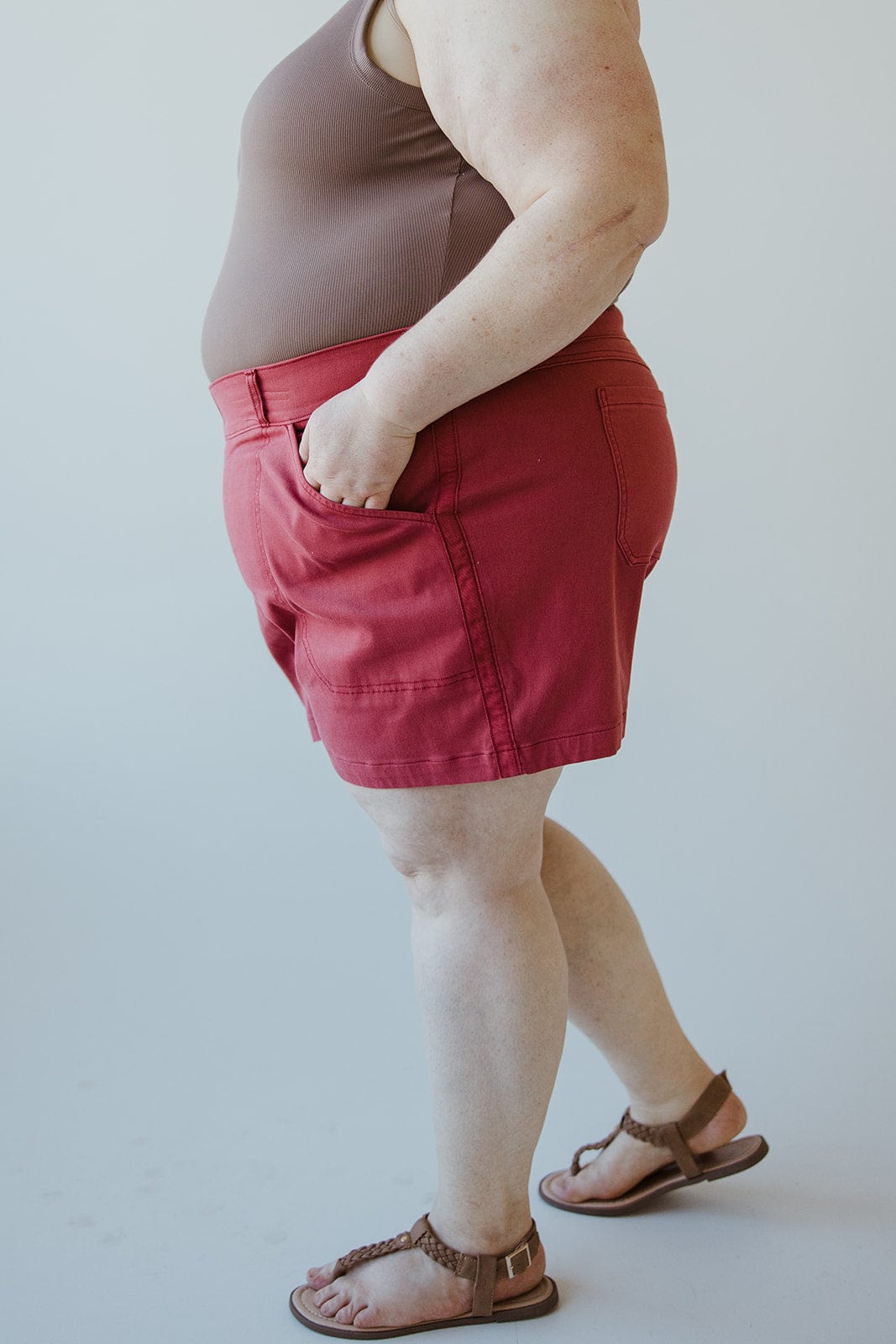 Person in a brown sleeveless top and Spanx® STRETCH TWILL SHORTS 6" IN WILD ROSE, with hands in pockets, standing against a plain background.