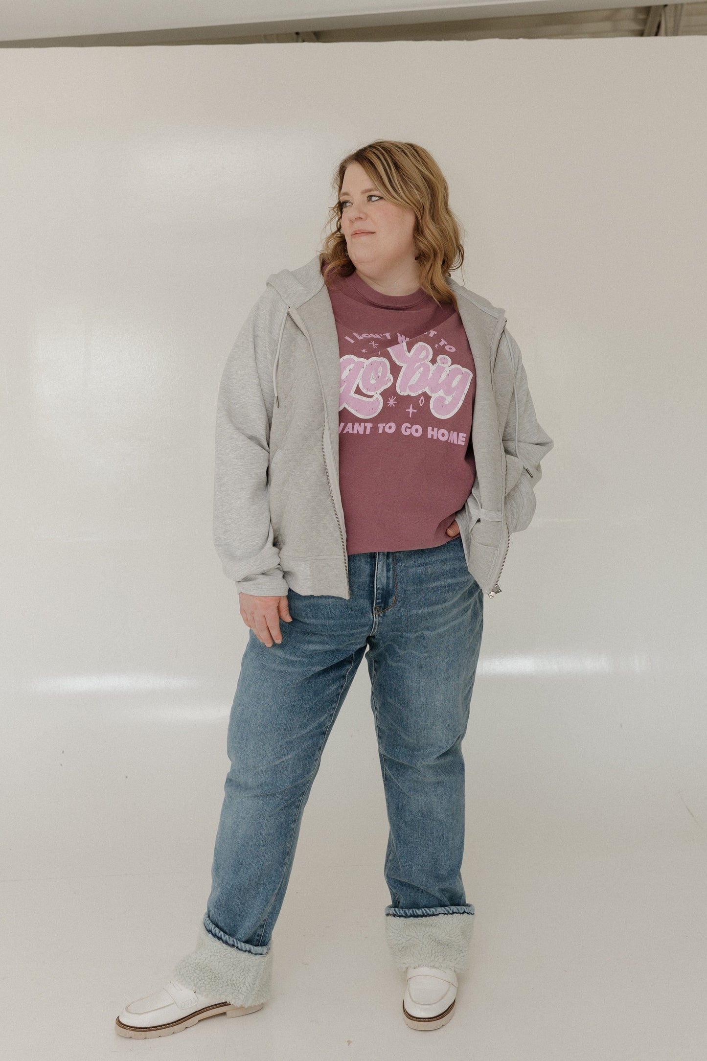Person wearing a gray jacket, berry graphic t-shirt, blue jeans, and white shoes on a plain background