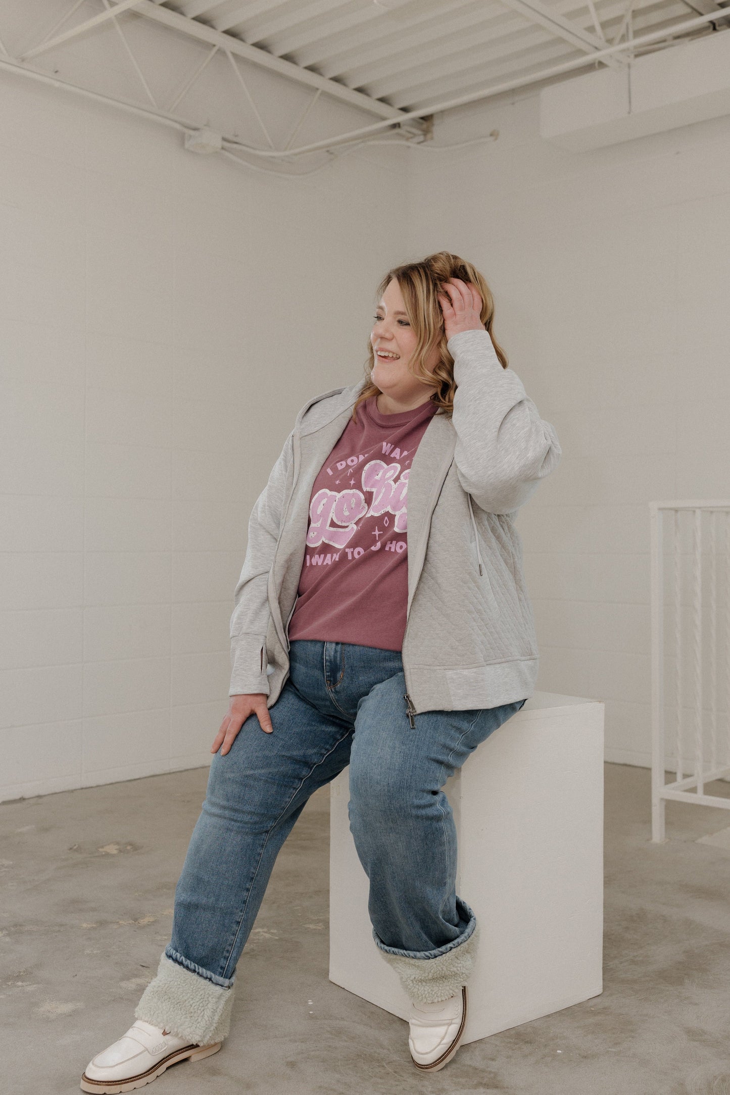 Person sitting on a white block in a minimalistic room wearing a gray jacket, berry graphic t-shirt, blue jeans, and white shoes.