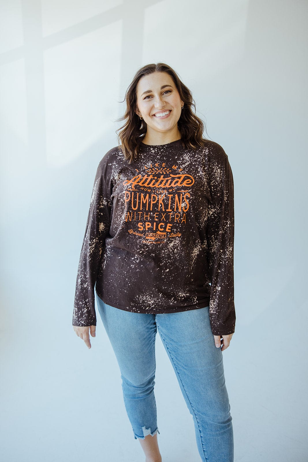A person smiles while wearing Love Marlow's hand-bleached "I LIKE MY ATTITUDE HOW I LIKE MY PUMPKINS WITH EXTRA SPICE" long-sleeve tee and light blue jeans against a light background.