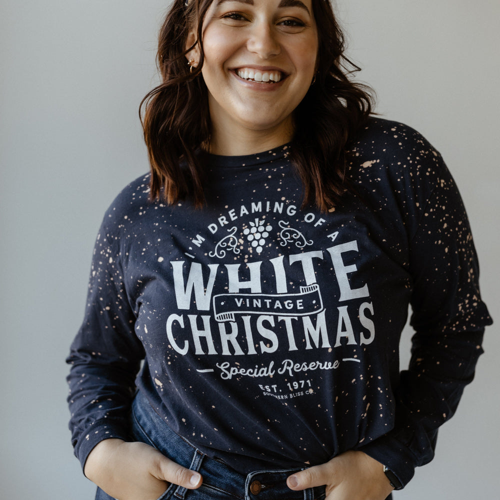 Person smiling, wearing a hand bleached dark speckled sweater with "I'm Dreaming of a White Christmas" print and jeans, perfectly capturing the holiday season vibe, standing against a plain background.