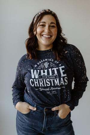 Person smiling, wearing a hand bleached dark speckled sweater with "I'm Dreaming of a White Christmas" print and jeans, perfectly capturing the holiday season vibe, standing against a plain background.