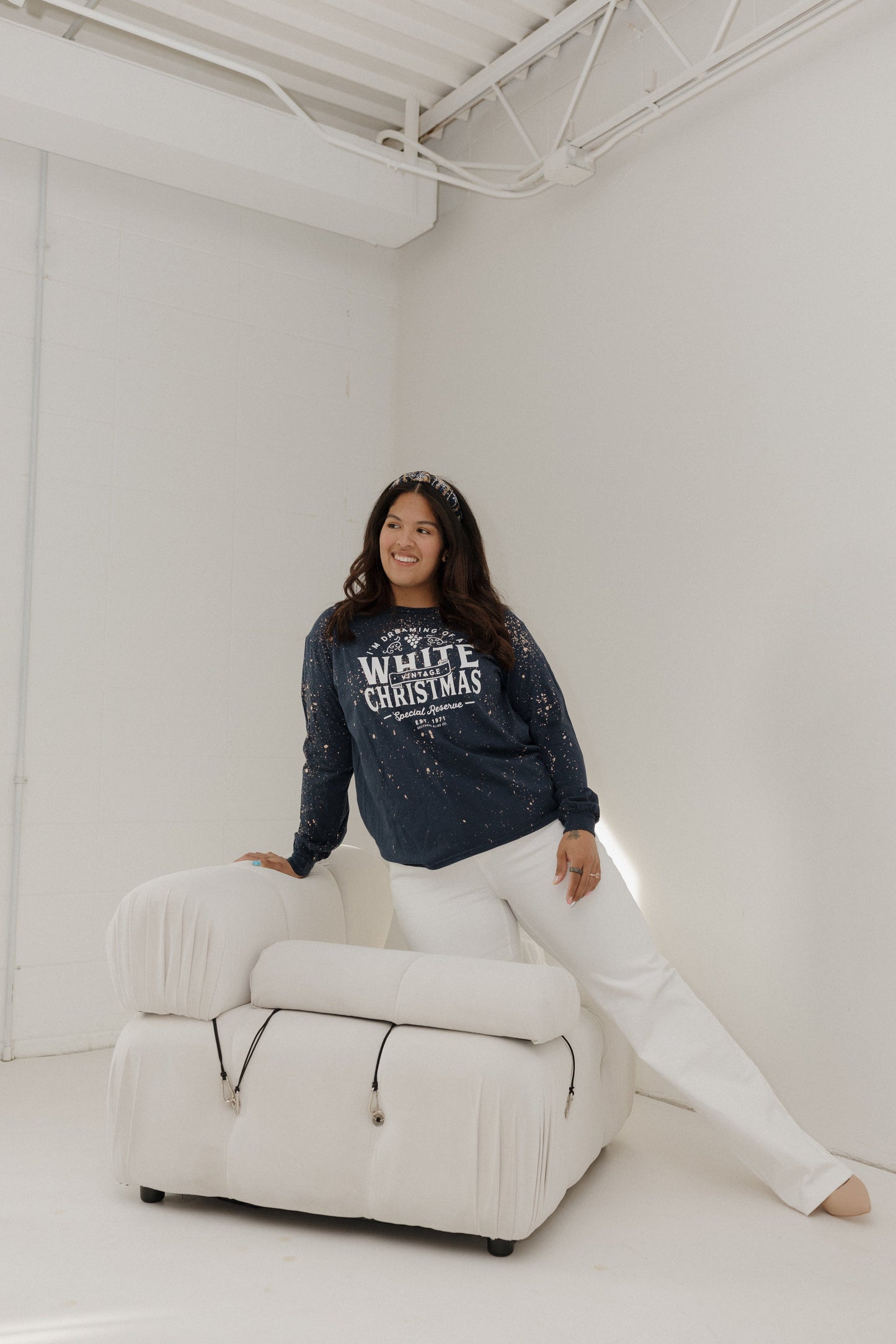Woman wearing a navy sweater with text sitting on a white chair in a minimal white room.