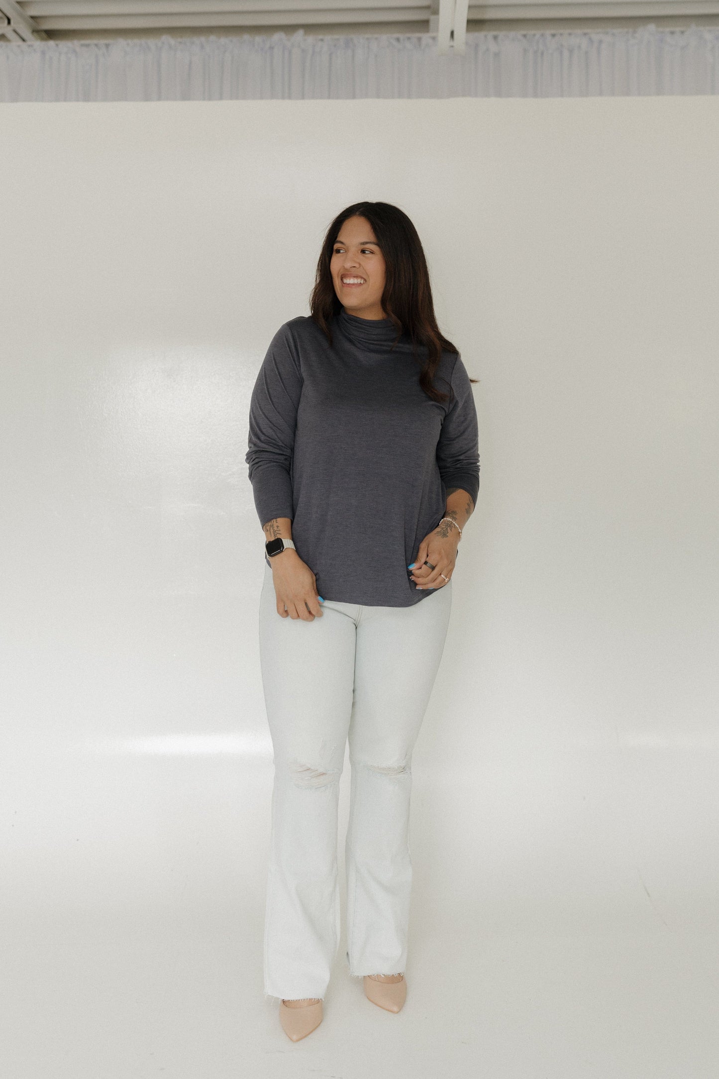 Woman wearing a gray turtleneck and white pants standing against a plain white wall.