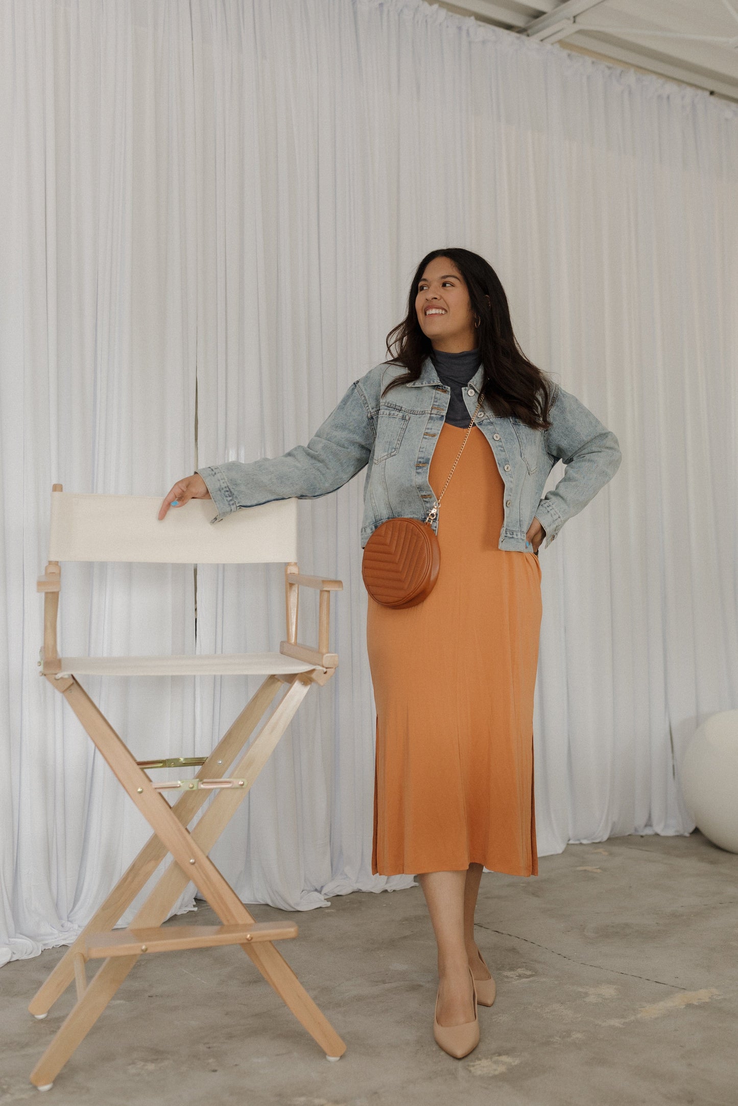 Woman in an orange dress and denim jacket standing next to a director's chair against a white curtain.