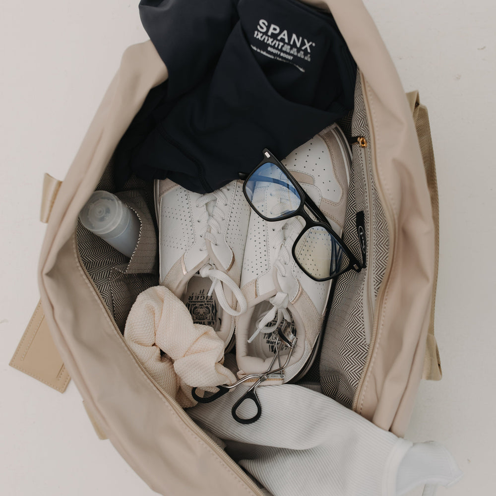 Open beige bag with black clothing, sneakers, and glasses on a white background