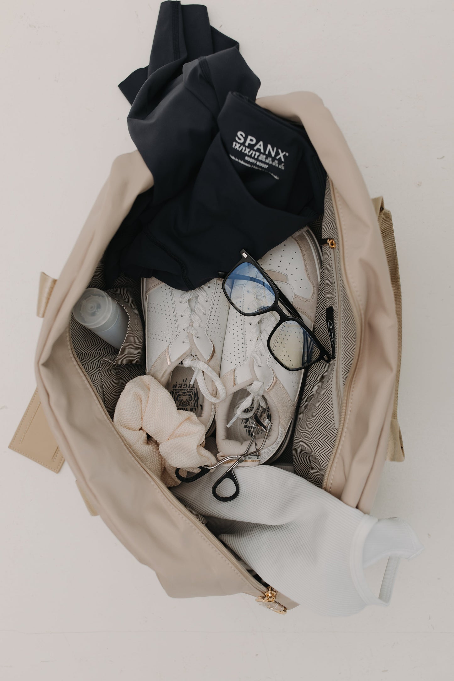 Open beige bag with black clothing, sneakers, and glasses on a white background
