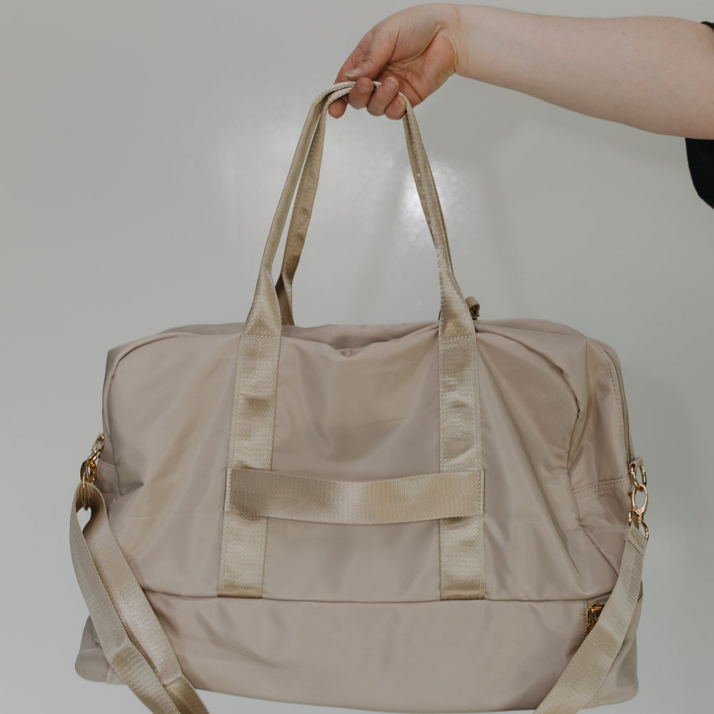 Beige duffel bag held by a hand against a plain background