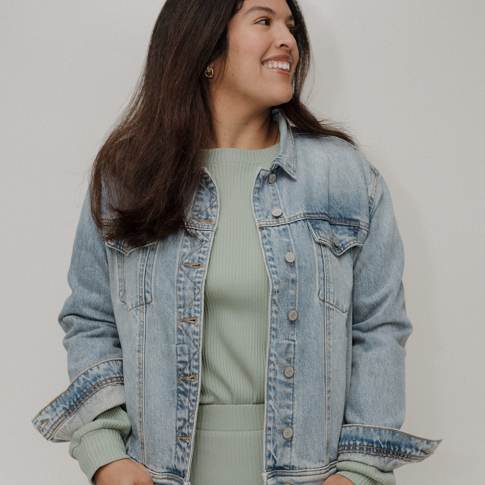Woman wearing a light blue denim jacket over a green top and skirt on a plain background
