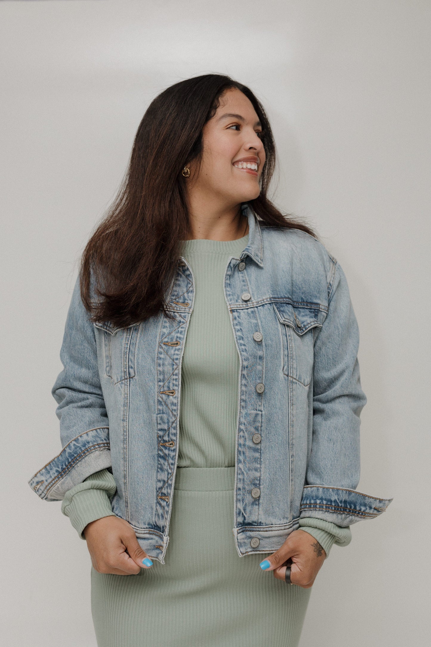 Woman wearing a light blue denim jacket over a green top and skirt on a plain background