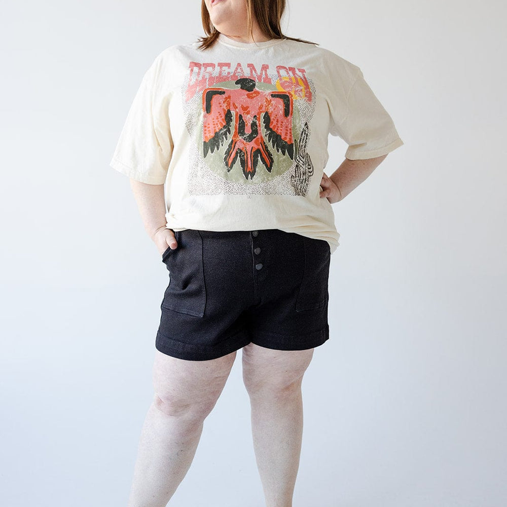 A woman stands against a plain background, wearing a graphic T-shirt, Judy Blue JUDY BLUE BUTTON FLY SHORTS WITH UTILITY POCKETS IN BLACK, and brown sandals. She has her hand on her hip and is looking slightly to the side, radiating cool 90s vibes.