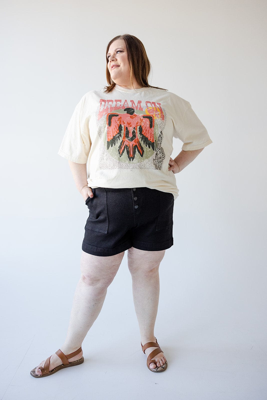 A woman stands against a plain background, wearing a graphic T-shirt, Judy Blue JUDY BLUE BUTTON FLY SHORTS WITH UTILITY POCKETS IN BLACK, and brown sandals. She has her hand on her hip and is looking slightly to the side, radiating cool 90s vibes.