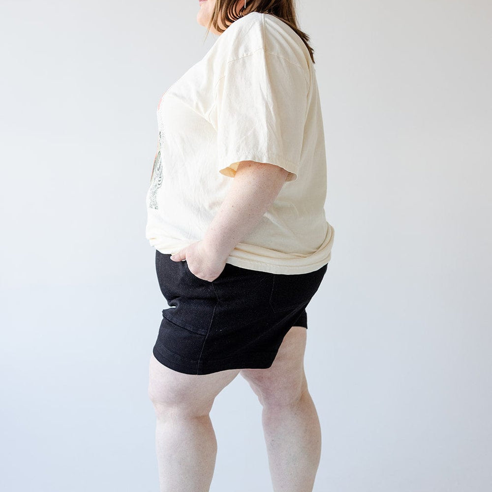 A person standing sideways with hands in pockets, wearing a white t-shirt, JUDY BLUE BUTTON FLY SHORTS WITH UTILITY POCKETS IN BLACK by Judy Blue, and brown sandals.