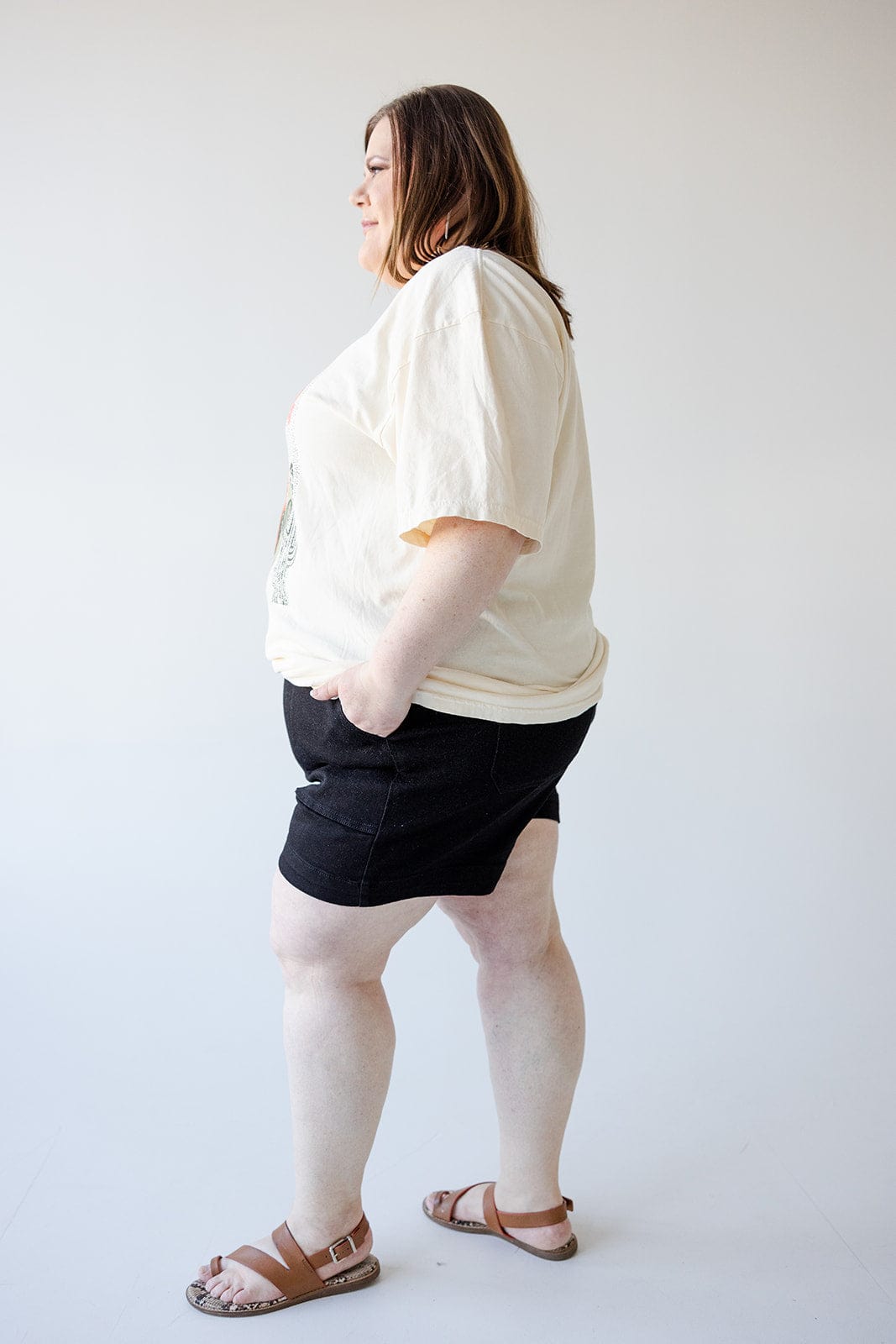 A person standing sideways with hands in pockets, wearing a white t-shirt, JUDY BLUE BUTTON FLY SHORTS WITH UTILITY POCKETS IN BLACK by Judy Blue, and brown sandals.