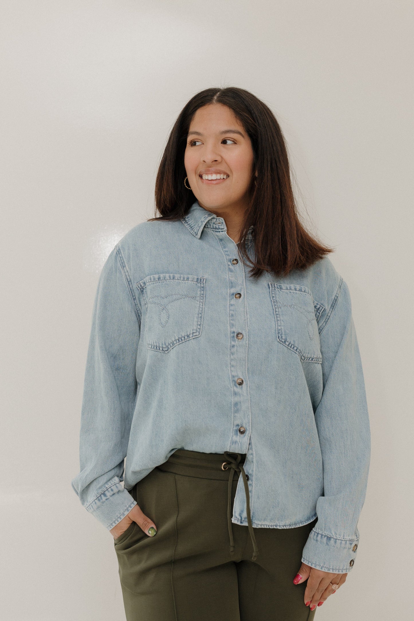 Woman wearing a light blue denim jacket and green pants against a plain background