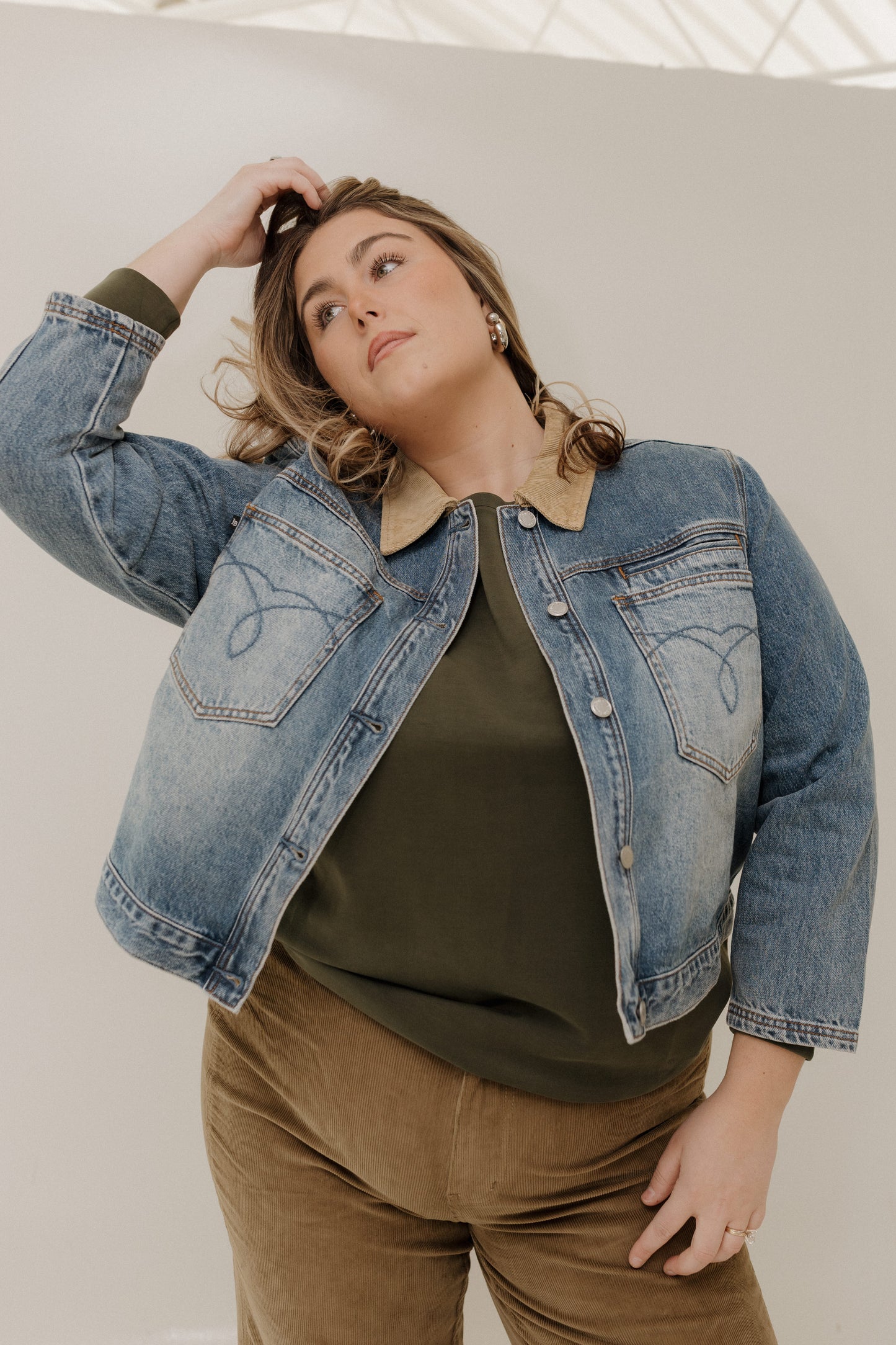 Woman wearing a denim jacket over a green top and brown pants against a white background