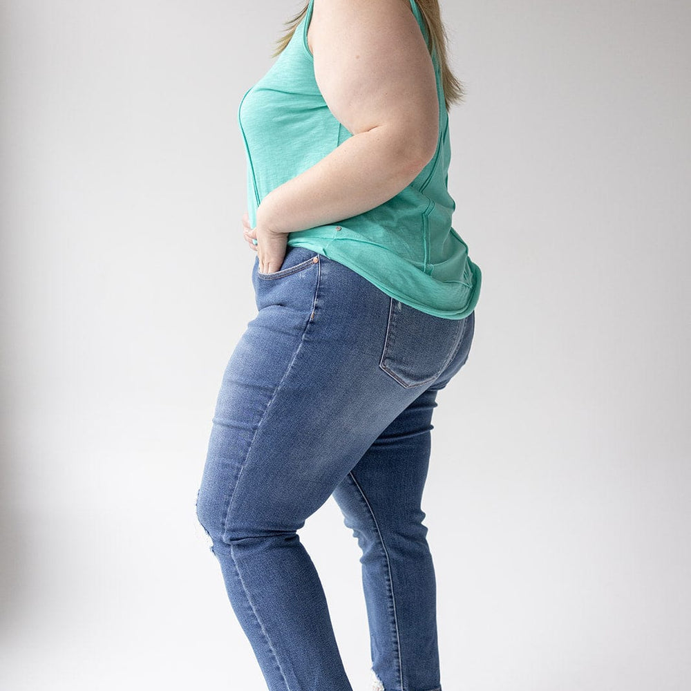 A woman stands in side profile, wearing a turquoise tank top, JUDY BLUE EMBROIDERED DESTROYED BOYFRIEND JEANS from Judy Blue, and red sneakers against a plain white background.