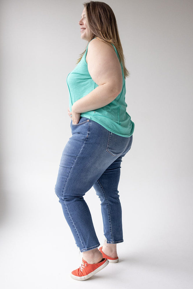 A woman stands in side profile, wearing a turquoise tank top, JUDY BLUE EMBROIDERED DESTROYED BOYFRIEND JEANS from Judy Blue, and red sneakers against a plain white background.