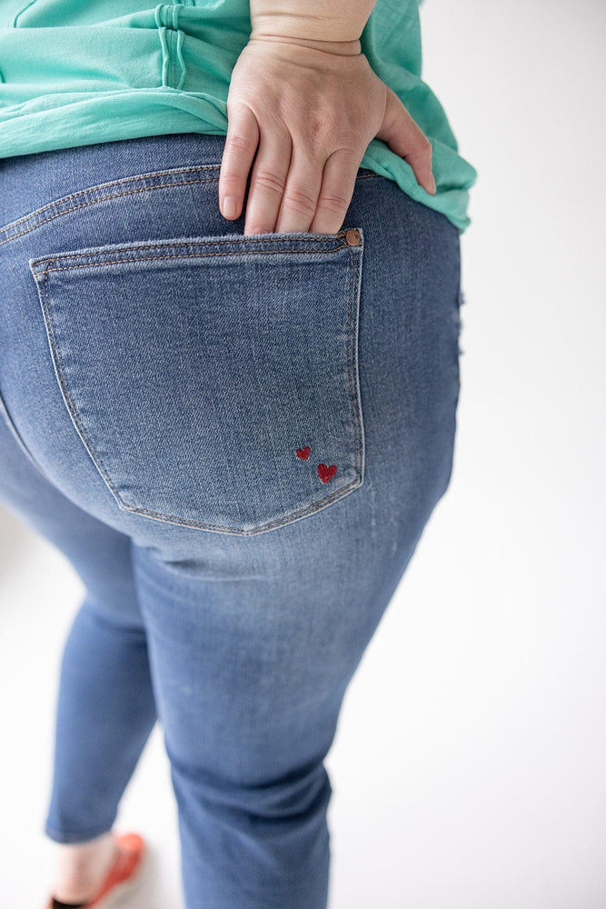 A person casually touching their back pocket with one hand while wearing the Judy Blue Embroidered Destroyed Boyfriend Jeans, adorned with small red heart designs on the pocket.