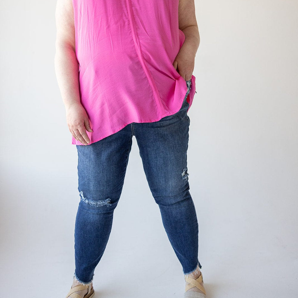 A person wearing a pink sleeveless top and Judy Blue distressed skinny jeans with beige sandals stands against a plain background.