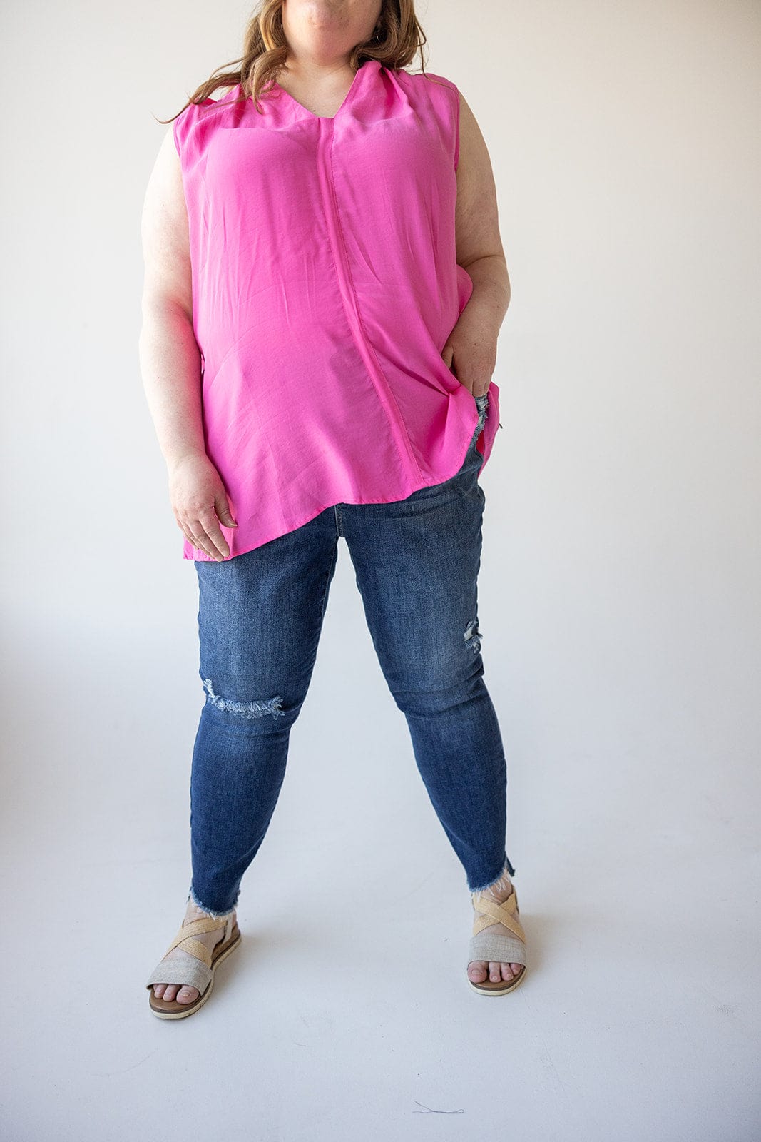 A person wearing a pink sleeveless top and Judy Blue distressed skinny jeans with beige sandals stands against a plain background.