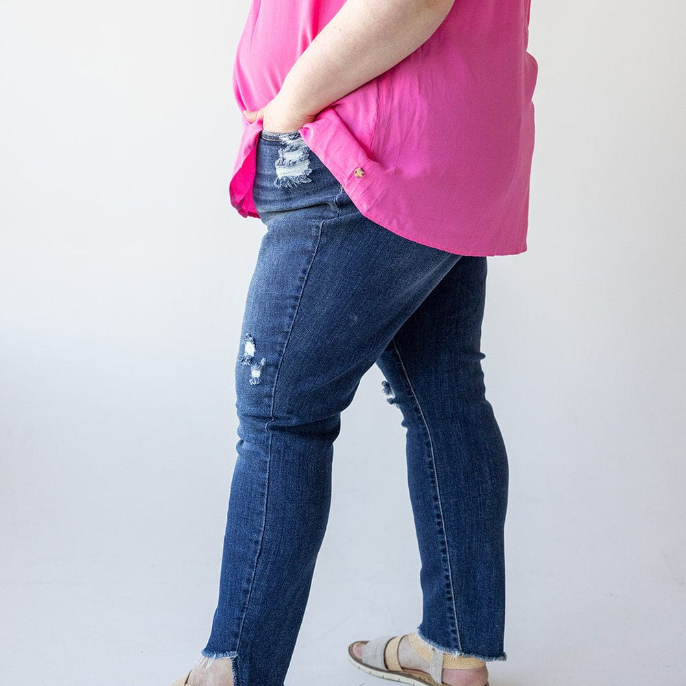 A person wearing a bright pink sleeveless top and distressed Judy Blue skinny jeans, paired with beige sandals, stands in a side view with hands in pockets against a plain white background.