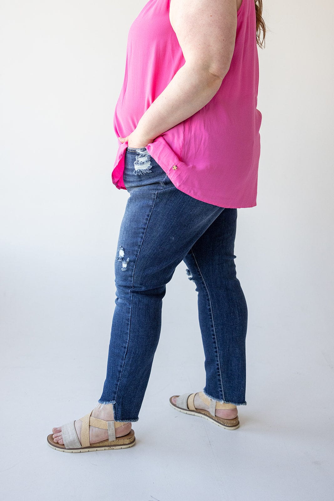 A person wearing a bright pink sleeveless top and distressed Judy Blue skinny jeans, paired with beige sandals, stands in a side view with hands in pockets against a plain white background.