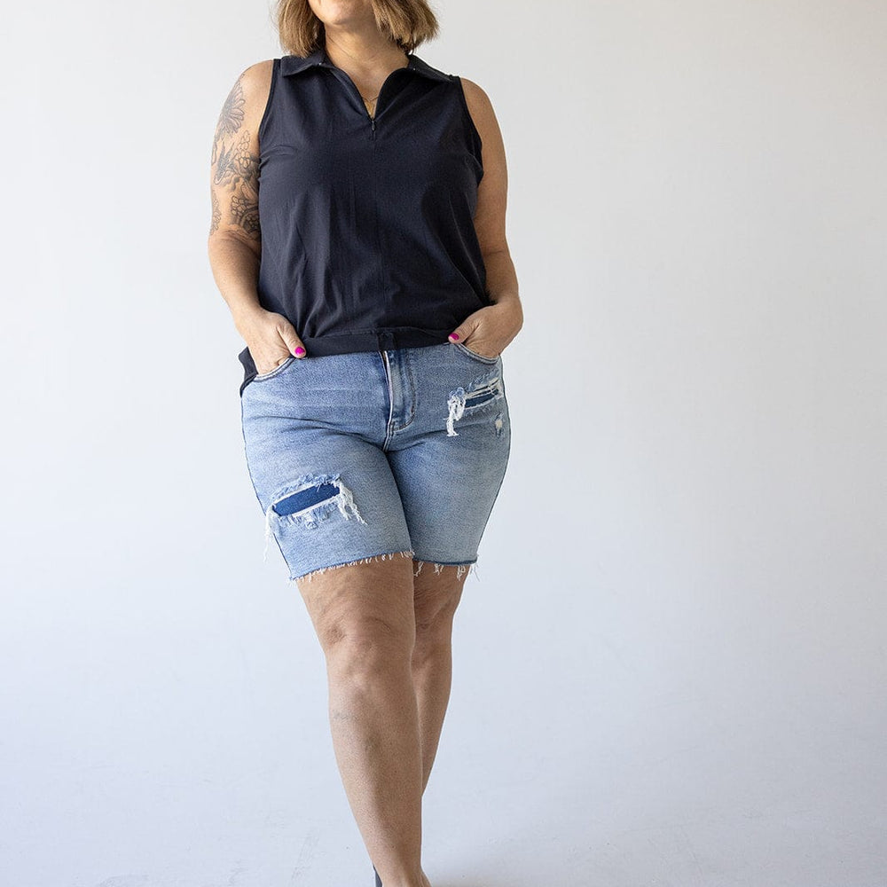 A woman in a black sleeveless top and plus-size denim shorts with a high-rise waistline stands against a plain background, looking to the side with hands in pockets.